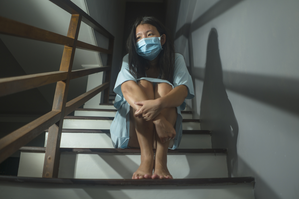 young scared and worried Asian Korean woman in protective mask sitting on stairs at home lockdown and quarantine during covid-19 virus pandemic in dramatic light