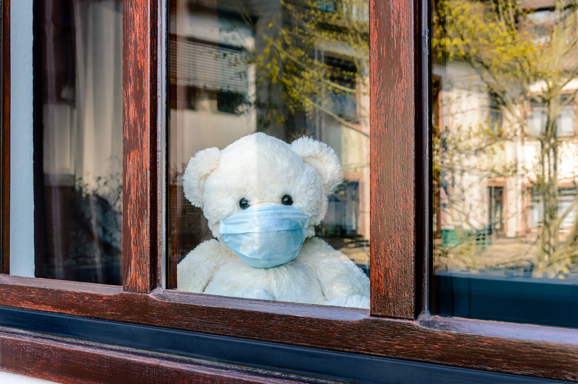 Teddy bear with protective medical mask looks out of the window on quarantine. Stay home concept